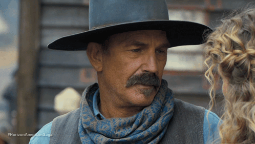 A Man With A Mustache Wearing A Cowboy Hat And Scarf Talks To A Woman In A Horizon American Saga Ad GIF