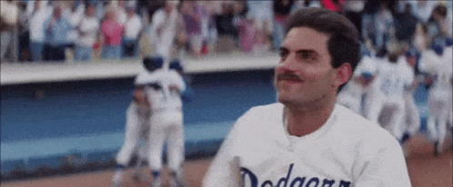 A Man With A Mustache Wearing A Dodgers Jersey Is Standing On A Baseball Field . GIF