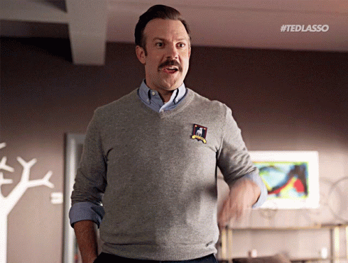 A Man With A Mustache Wearing A Grey Sweater With A Patch That Says " Ted Lasso " GIF