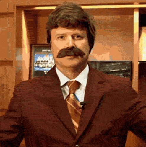 A Man With A Mustache Wearing A Suit And Tie Stands In Front Of A Television GIF