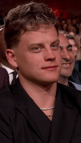 A Man With A Necklace Around His Neck Is Sitting In A Crowd Of People GIF