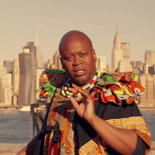 A Man With A Necklace Around His Neck Stands In Front Of A City GIF