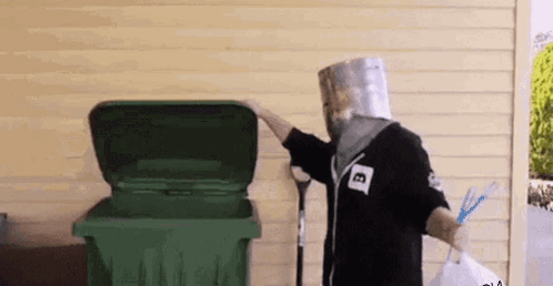 A Man With A Paper Bag On His Head Is Standing Next To A Green Trash Can . GIF