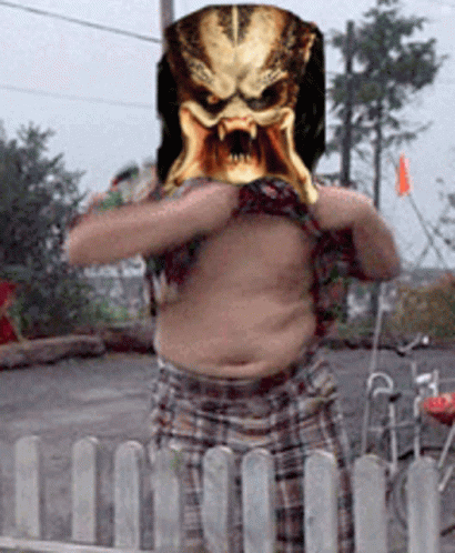 A Man With A Predator Mask On His Head Stands In Front Of A Fence GIF