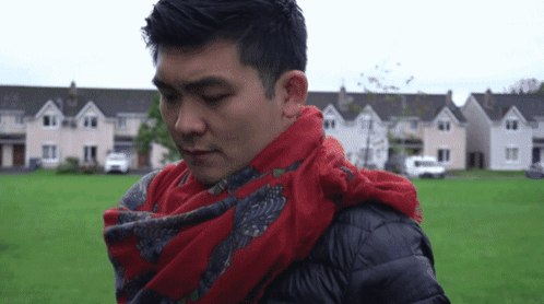 A Man With A Red Scarf Around His Neck Is Standing In Front Of A Row Of Houses GIF