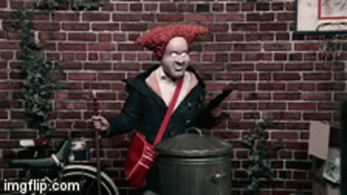 A Man With A Red Wig Is Standing In Front Of A Brick Wall Holding A Trash Can GIF