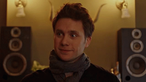 A Man With A Scarf Around His Neck Is Smiling In Front Of A Pair Of Speakers GIF