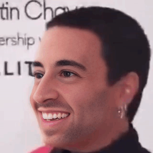 A Man With A Shaved Head And Earrings Is Smiling In Front Of A Sign That Says Tin Chow . GIF