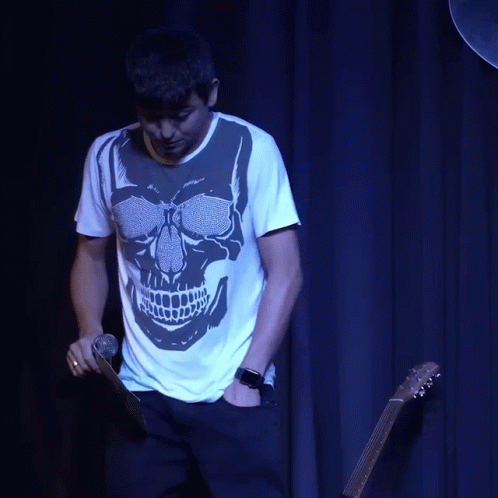 A Man With A Skull On His Shirt Is Holding A Guitar And Talking On A Cell Phone GIF