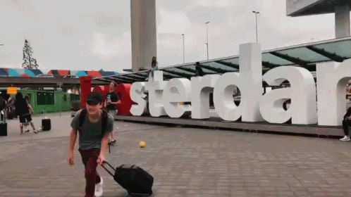 A Man With A Suitcase Is Walking In Front Of A Large Amsterdam Sign GIF