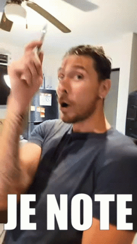 A Man With A Surprised Look On His Face Is Holding A Cell Phone In Front Of A Sign That Reads Je Note GIF