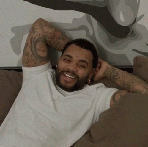 A Man With A Tattoo On His Arm Is Laying On A Couch And Smiling GIF