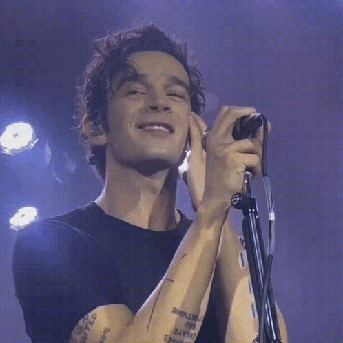 A Man With A Tattoo On His Arm Is Singing Into A Microphone While Smiling GIF
