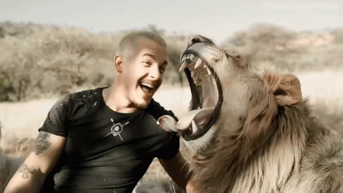 A Man With A Tattoo On His Arm Is Sitting Next To A Lion With Its Mouth Wide Open GIF