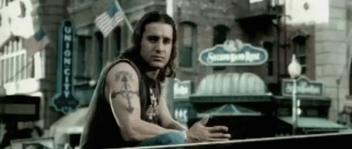 A Man With A Tattoo On His Arm Is Sitting On A Railing In Front Of A Building With A Sign That Says Second Base GIF