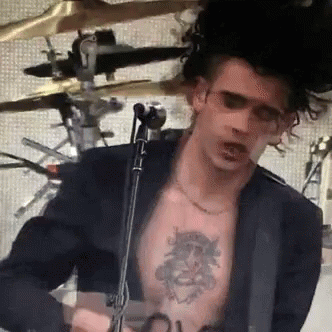 A Man With A Tattoo On His Chest Is Playing A Guitar In Front Of A Microphone GIF