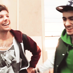 A Man With A Tattoo On His Chest Is Smiling Next To Another Man Wearing A Green Vest GIF