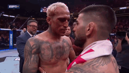 A Man With A Tattoo On His Chest Stands Next To Another Man With A Bud Light Banner Behind Him GIF