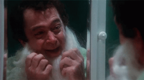 A Man With A White Beard Is Looking At Himself In A Mirror . GIF