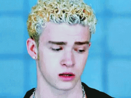 A Man With Blonde Curly Hair Is Wearing A Black Shirt And A Chain Around His Neck . GIF