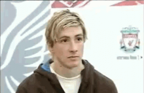 A Man With Blonde Hair And A Brown Jacket Is Standing In Front Of A Liverpool Logo . GIF