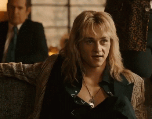 A Man With Blonde Hair And A Necklace Is Sitting On A Couch In Front Of A Man In A Suit . GIF