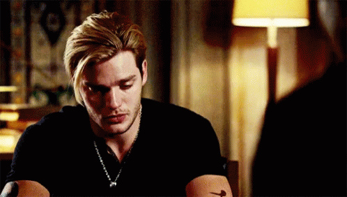 A Man With Blonde Hair And A Tattoo On His Arm Is Sitting At A Table With A Lamp In The Background . GIF