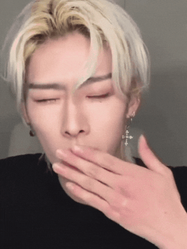 A Man With Blonde Hair And Earrings Is Covering His Mouth With His Hand GIF