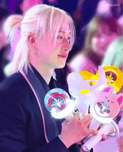 A Man With Blonde Hair Is Holding A Light Stick And A Stuffed Animal GIF