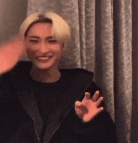 A Man With Blonde Hair Is Smiling And Waving At The Camera While Wearing A Black Jacket . GIF