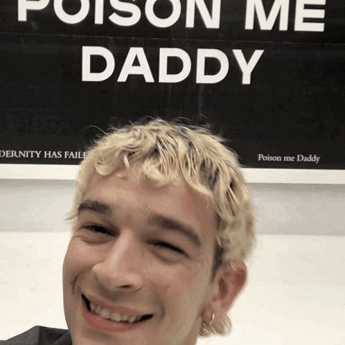 A Man With Blonde Hair Is Smiling In Front Of A Sign That Says Poison Me Daddy GIF