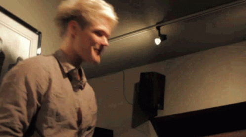 A Man With Blonde Hair Is Standing In A Room With A Picture On The Wall Behind Him GIF