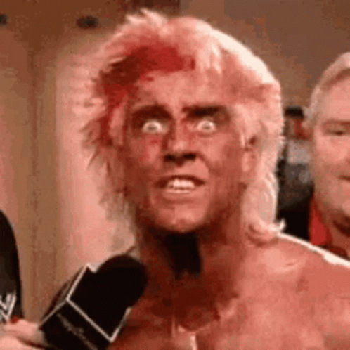 A Man With Blood On His Face Is Talking Into A Microphone While Wearing A Mullet . GIF