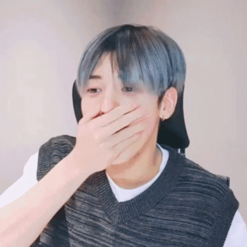 A Man With Blue Hair Is Covering His Mouth With His Hand While Wearing A Sweater Vest . GIF