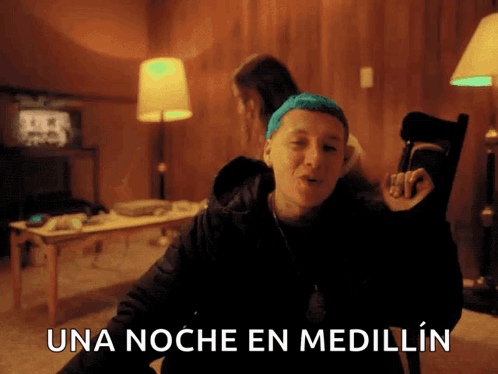 A Man With Blue Hair Is Sitting In A Chair With The Words Una Noche En Medellin Above Him GIF