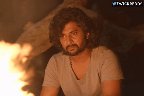 A Man With Curly Hair And A Beard Is Sitting In Front Of A Fire With The Hashtag 7Wickreddy Above Him GIF