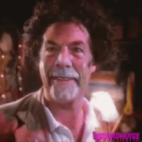 A Man With Curly Hair And A Beard Is Smiling And Looking At The Camera . GIF