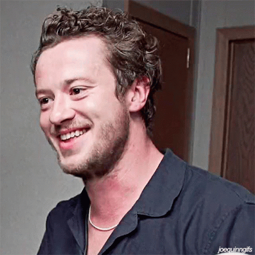 A Man With Curly Hair And A Beard Is Smiling And Wearing A Necklace . GIF