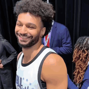 A Man With Curly Hair And A Beard Is Smiling For The Camera While Wearing A Basketball Jersey . GIF