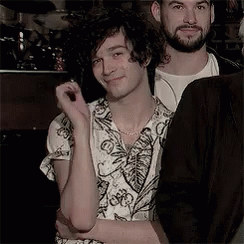 A Man With Curly Hair And A Beard Is Standing Next To Another Man In A Room . GIF