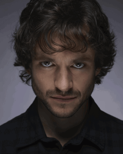 A Man With Curly Hair And A Beard Looks At The Camera With A Serious Look On His Face GIF