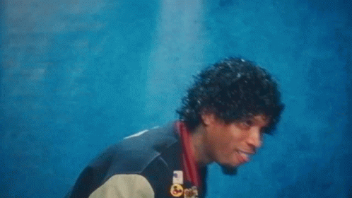 A Man With Curly Hair And A Varsity Jacket Is Standing In Front Of A Blue Wall . GIF