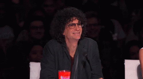 A Man With Curly Hair And Glasses Is Sitting In Front Of A Microphone Holding A Cup Of Coca Cola . GIF