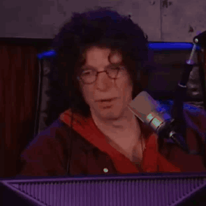 A Man With Curly Hair And Glasses Is Sitting In Front Of A Microphone In Front Of A Computer Monitor . GIF