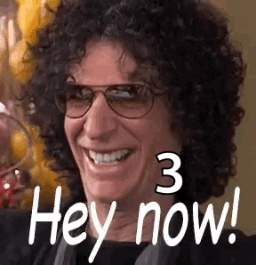 A Man With Curly Hair And Glasses Is Smiling And Saying Hey Now . GIF