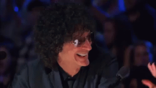 A Man With Curly Hair And Sunglasses Is Smiling In Front Of A Crowd In A Dark Room . GIF