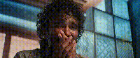 A Man With Curly Hair Is Covering His Mouth With His Hands In A Room . GIF