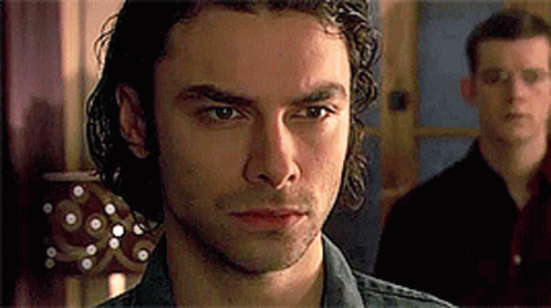 A Man With Curly Hair Is Looking At The Camera While Another Man Stands Behind Him . GIF