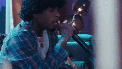 A Man With Curly Hair Is Singing Into A Microphone While Sitting On A Couch GIF