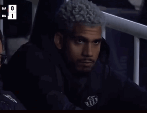 A Man With Curly Hair Is Sitting In A Stadium Watching A Game With The Score Bar 0 Leg 1 GIF
